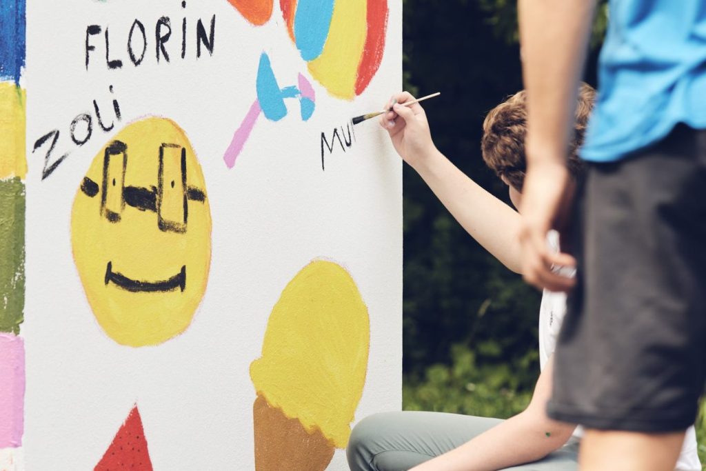 Ein Schüler bemalt mit Farbe und Pinsel eine weiße Wand auf der bereits andere Symbole wie ein Eis, Schmetterling und Smiley gemalt ist.