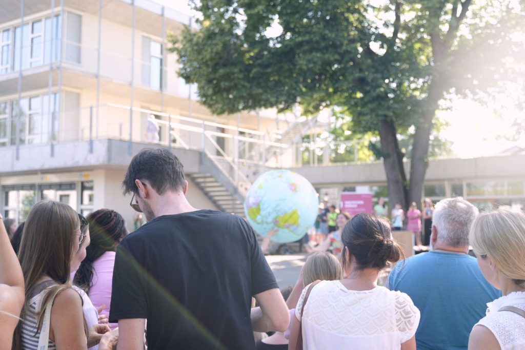 Zu sehen ist ein Schulhof mit Eltern, Lehrer*innen und Schüler*innen. Im Hintergrund ist ein Baum und ein Schulgebäude zu erkennen. Im Zentrum schwebt eine Weltkugel.