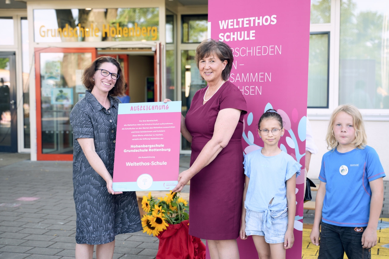 Zwei Frauen lächeln in die Kamera und halten gemeinsam eine Urkunde in den Händen. Neben ihnen stehen ein Junge und ein Mädchen. Auf der Urkunde steht: Auszeichnung Hohenbergschule Grundschule Rottenburg Weltethos Schule.