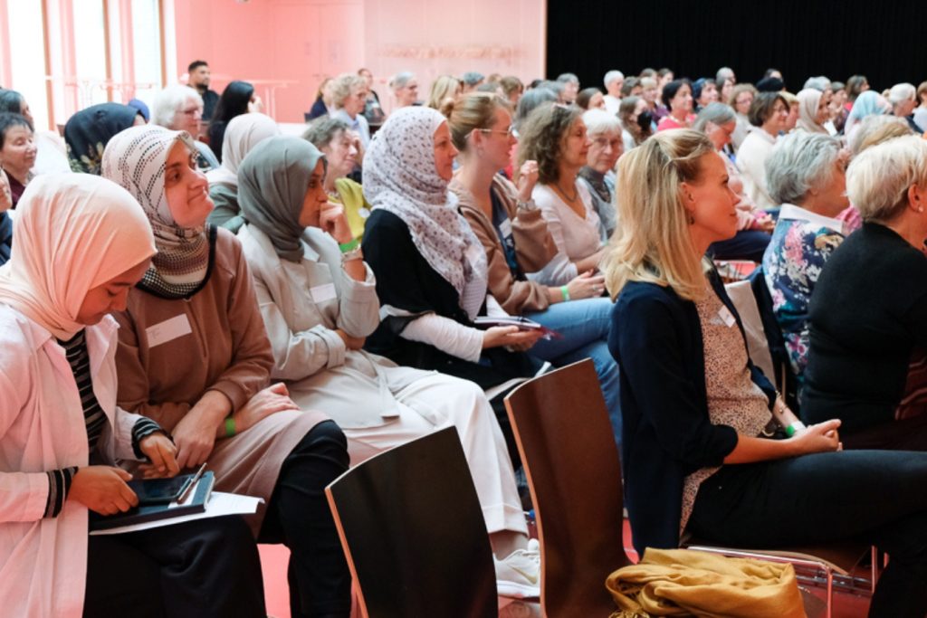 Viele unterschiedliche Frauen sitzen in einem großen Raum auf Stühlen und hören konzentriert zu.