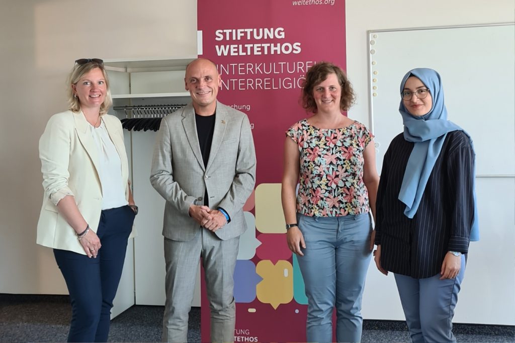 Drei Frauen und ein Mann stehen vor dem Roll-up der Stiftung Weltethos und blicken lächelnd in die Kamera.